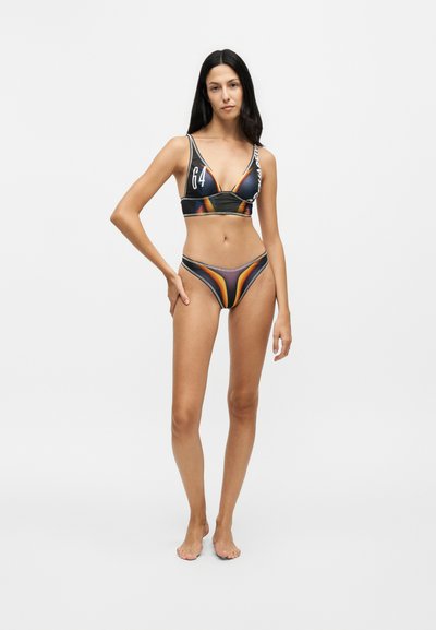 Femme debout pieds nus portant un bikini à motifs noirs, orange et gris avec le numéro 64 sur le haut sur fond blanc.