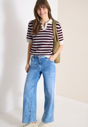 Kvinde smilende iført en stribet kortærmet sweater, lyseblå vide jeans, beige sneakers og bærer en olivengrøn skuldertaske.