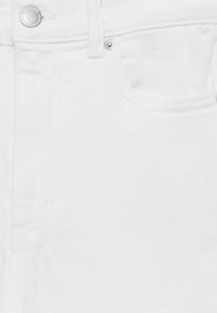 Detail bílého denimového materiálu, který ukazuje přední kapsu, poutko na pásek a dvě kovové nýty s vyraženým logem značky.