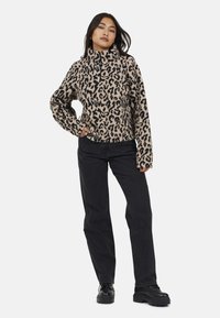 Donna in piedi con la mano in tasca, che indossa un pullover fuzzy beige e nero con stampa leopardata, jeans neri a gamba dritta e scarpe chunky nere.