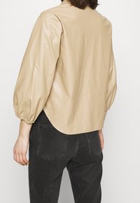 Beige konstläderblus med ballongsärmar, rundat nederkant och slät textur, kombinerad med mörka denimjeans.