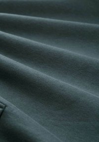 Ciemnozielony materiał wyeksponowany w delikatnych fałdach, charakteryzujący się gładką teksturą i subtelnym wzorem tkanym. Miękki materiał z lekkim połyskiem.