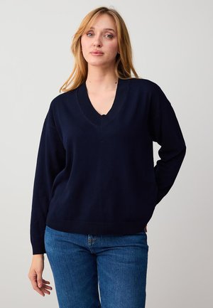 Donna con capelli biondi lunghi che indossa un maglione blu navy a scollo a V e jeans blu, in piedi davanti a uno sfondo chiaro e uniforme.