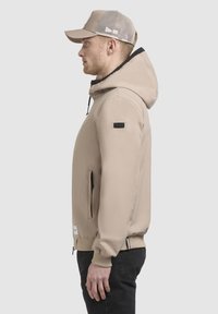 Giacca beige con cappuccio e dettagli neri, tessuto liscio, polsini elasticizzati e tasche anteriori con zip. Indossata con un cappellino beige in rete.