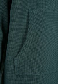 Tessuto a nido d'ape verde con tasca frontale, superficie testurizzata e dettagli di cucitura a contrasto, che offre un design accogliente e funzionale.
