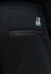 Μαύρο ύφασμα με οριζόντια τσέπη με φερμουάρ και λογότυπο K-Way σε μπλε, λευκό και κόκκινο πάνω από την τσέπη στην πάνω δεξιά πλευρά.