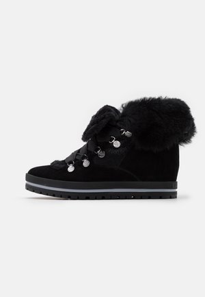 Bottine noire avec semelle épaisse en caoutchouc, col et languette en fausse fourrure, œillets métalliques et larges lacets ruban sur fond blanc.