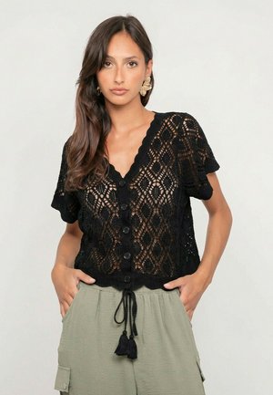 Femme portant un haut noir en crochet boutonné avec des bords festonnés et un pantalon à cordon vert sauge, debout les mains dans les poches.