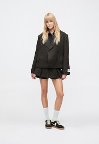 Fiatal nő hosszú szőkés hajjal, sötét, túlméretezett blézert és pliszírozott szoknyát visel, fehér zokniban, fekete-fehér sportcipőben áll.