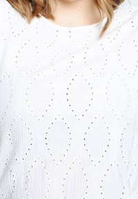 Haut en tissu blanc texturé avec des motifs d'œillets complexes porté par une personne aux cheveux châtain clair mi-longs.