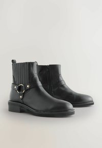 Botines de cuero negro hasta el tobillo con un panel acanalado, cremallera lateral y una correa decorativa con acento en hardware de color dorado. Suela plana.