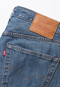 Denim jeans in medium blue with a tan leather patch on the waistband, featuring orange stitching and a red Levi's tag on the back pocket.