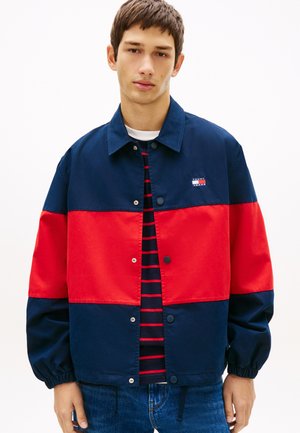 Ung mand iført en navy og rød farveblokeret jakke over en navy og rødstribet skjorte, stående foran en ensfarvet hvid baggrund.