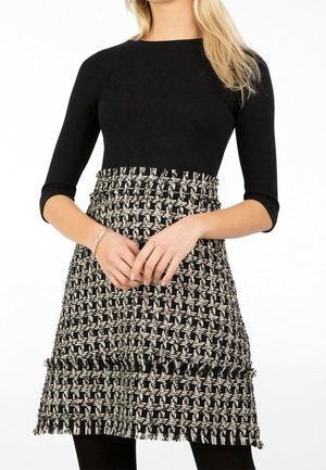 Femme portant un haut noir à manches trois-quarts et une jupe taille haute en tweed noir et blanc avec des détails en franges.