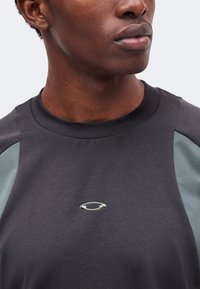 Jeune personne à la peau foncée portant une chemise gris foncé avec des panneaux gris clair et un petit logo ovale sur la poitrine.