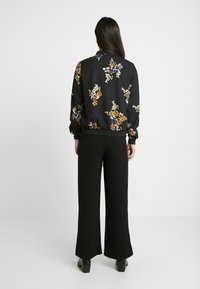 Black bomber jacket with colorful floral print, elastic cuffs, and hem. Paired with wide-leg black trousers and heeled boots.
