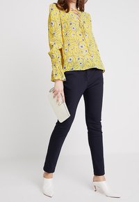 Blouse florale jaune avec manches évasées, associée à un pantalon slim-fit bleu marine. Les accessoires comprennent un sac à main blanc et des chaussures à talons pointus blanches.