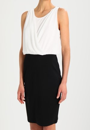 Ärmelloses Kleid mit weißem, drapiertem Oberteil und einem enganliegenden schwarzen knielangen Rock an einer Person vor schlichtem Hintergrund.