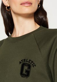 Donna che indossa una felpa verde oliva con l'emblema nero "ATHLETIC GAP DEPT.", orecchini a cerchio dorati e lunghi capelli scuri.