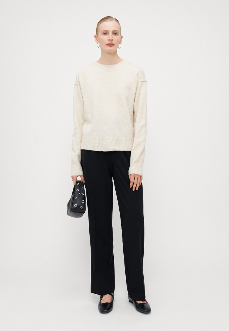 Pull en maille blanc cassé à manches longues, associé à un pantalon noir ; portant un sac noir avec des accents de rivets en métal ; chaussures élégantes.