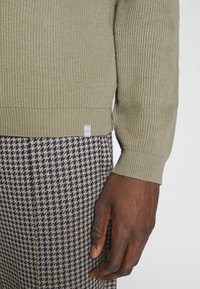 Sweter w kolorze oliwkowej zieleni z prążkowaną teksturą i mankietami; uzupełniony spodniami w kratę houndstooth w odcieniach granatu i beżu.