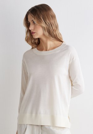 Kvinde med bølget lysebrunt hår iført en cremefarvet langærmet sweater, der kigger til venstre mod en ensartet hvid baggrund.