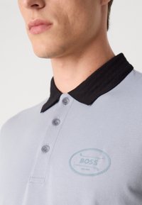 Lys grå polo skjorte med svart ribbestrikket krage, tre knappeåpninger og et brodert blått "BOSS"-logo på brystet.