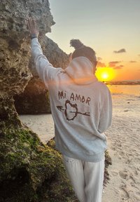 Person mit grauem "Mi Amor"-Hoodie lehnt während des Sonnenuntergangs über dem ruhigen Meer an einem moosbedeckten Felsen am Strand.