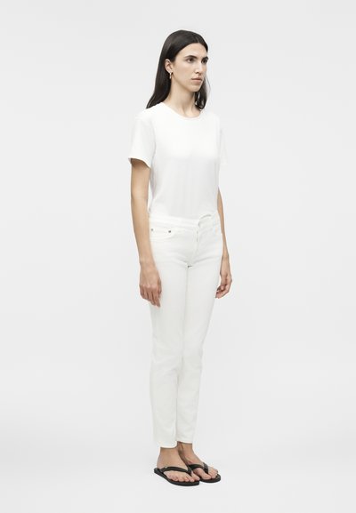 Femme debout portant une chemise blanche à manches courtes, un pantalon blanc, des tongs noires, avec de longs cheveux foncés, sur un fond clair uni.