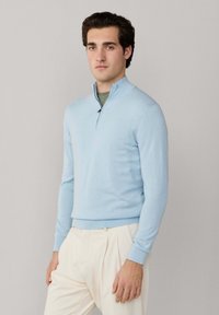 Jeune homme portant un pull à fermeture éclair courte bleu clair par-dessus une chemise verte et un pantalon plissé crème, debout devant un fond neutre.