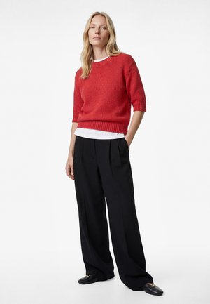 Blondynka stoi, ubrana w czerwony sweter z krótkim rękawem, białą podkoszulkę, szerokie czarne spodnie i czarne loafersy na białym tle.