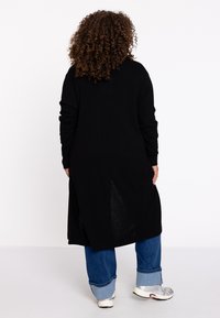 Schwarze Langarm-Cardigan mit lockerer Passform, versehen mit seitlichen Schlitzen und einem strukturierten Strickmuster. Kombiniert mit blauen, hochgekrempelten Jeans und silbernen Sneakern.