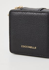 Musta teksturoidulla nahkalaukku, jossa on sileä pinta, varustettuna kultaisella vetoketjulla ja etupuolella kohokuvioidulla "Coccinelle"-logolla.