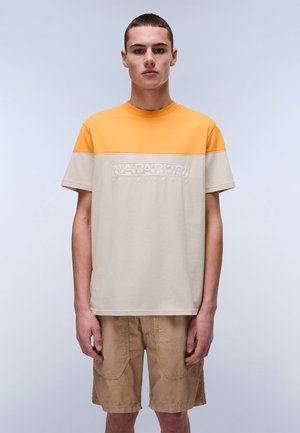 T-shirt of cotton blend, featuring orange upper and beige lower sections, with textured logo on chest. Model wears beige shorts.