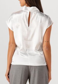 Weiße Satinbluse mit kurzen Ärmeln, hohem Ausschnitt und einem Schlüsselaugen-Ausschnitt hinten. Glatte Textur, lockere Passform, kombiniert mit grauen Hosen.