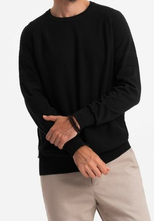 Mand iført sort ribstrikket sweater med beige bukser, justerer ærmet og bærer et brunt perlearmbånd på venstre håndled.