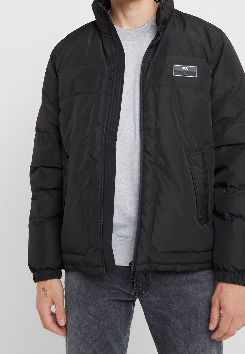 Veste matelassée noire avec fermeture éclair à l'avant, poignets élastiques et patch logo sur la poitrine. Présente une texture lisse et matelassée ainsi que des poches latérales.