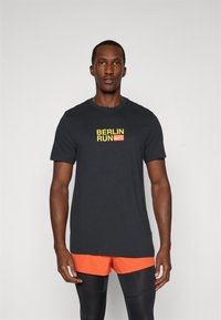 Black short-sleeve t-shirt with "BERLIN RUN" text in yellow and red. Worn with orange shorts and black leggings. Cotton material, casual fit.