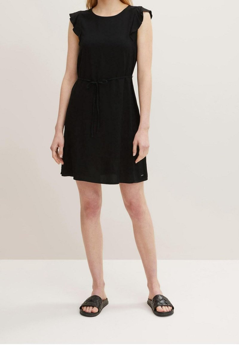 Robes sans manches noires avec des accents à volants, un col rond et une taille nouée. Portée avec des sandales à glissière noires sur un fond clair.