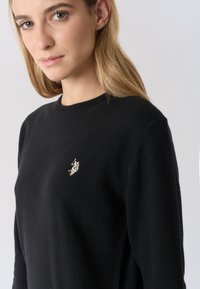 Maglione nero in maglia con scollo rotondo e piccolo logo argentato sul petto a sinistra. Morbida texture e vestibilità classica.