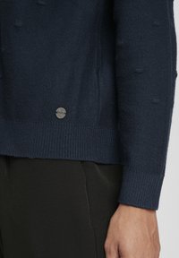 Marinblå stickad tröja med texturerat mönster, ribbad nederkant och ärmslut, med en liten metallpinne nära botten.