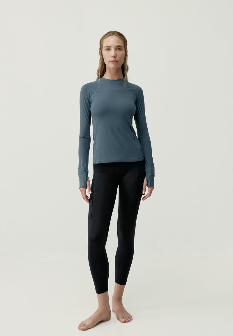 Dlhý rukáv športového trička v slate blue, skombinovaný s čiernymi, priliehavými legínami so bočnými vreckami. Hladká textúra, minimalistický dizajn.