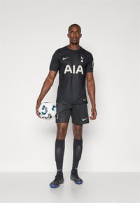 Tenue de football noire avec un design texturé, logo "AIA" en couleur claire et emblème de Tottenham Hotspur. Un joueur tient un ballon de football multicolore.