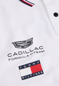Λευκό πόλο πουκάμισο με το λογότυπο της ομάδας Cadillac Formula 1 και το λογότυπο της σημαίας Tommy Hilfiger κάτω από το γιακά με ριγέ περιθώριο.