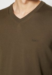 Nahaufnahme einer Person, die einen olivgrünen V-Ausschnitt-Pullover trägt, auf dessen linker Brust "BOSS" eingestickt ist.