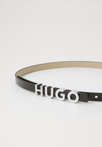 Cintura in pelle nera con design sottile e emblema in metallo argento "HUGO". Texture liscia con finitura lucida e chiusura con punzone regolabile.