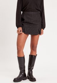 Schwarzer Minirock mit strukturierten Oberflächen und subtilen Glitzerpartikeln, mit einem seitlichen Schlitz. Kombiniert mit hohen schwarzen Lederstiefeln.