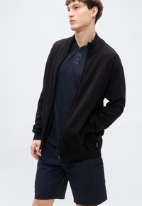 Μαύρο μπουφάν με φερμουάρ από μαλακό ύφασμα, με γιακά. Συνδυάζεται με μπλε κοντομάνικο τοπ και μπλε σορτς με λεπτομέρεια τσέπης.