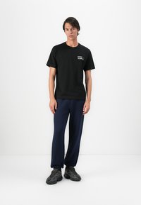 Jeune homme debout portant un t-shirt noir Lacoste, un pantalon de jogging bleu marine et des baskets noires devant un fond blanc uni.