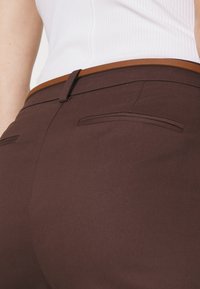 Pantalons marron sur mesure avec une texture lisse, présentant une coupe slim, des poches arrière discrètes et une ceinture contrastante beige.